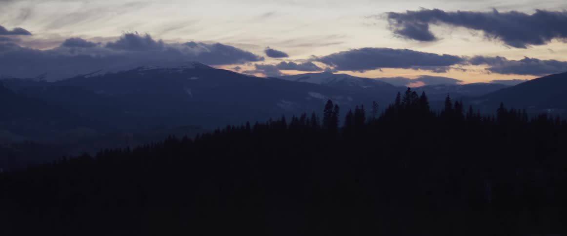 The last light of day fades behind a snowy mountain range, silhouetting a dense pine forest