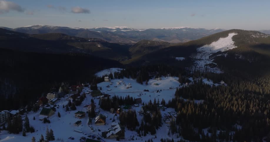 A static aerial view captures a quaint ski resort village nestled in a vast, snow-covered mountain range at sunset