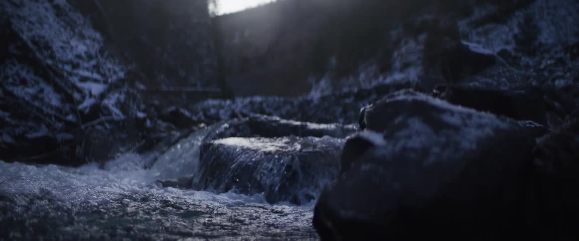 A slow-motion close-up captures a cold mountain stream flowing over snow-dusted rocks in a dim winter light