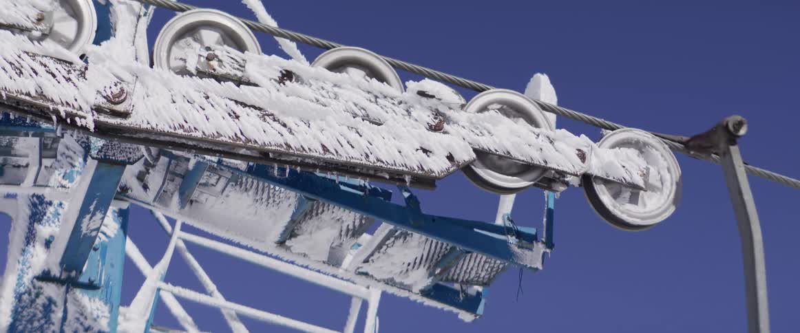Frozen ski lift pulley wheels slowly turn, covered in rime ice and snow, against a vibrant blue sky