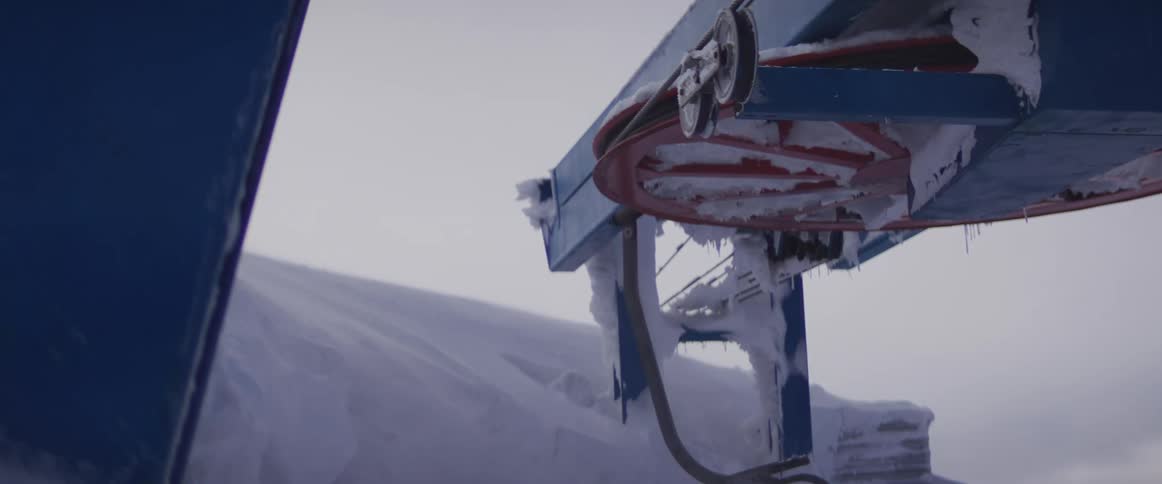 A close-up shot shows a frozen and snow-covered ski lift mechanism on a desolate winter day