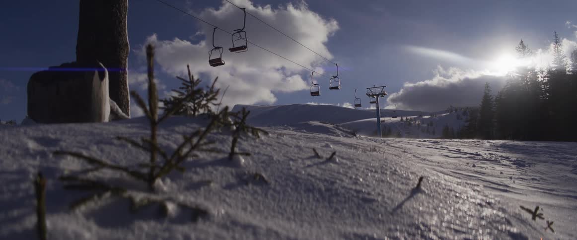 A strong wind blows fresh powder across a snowy mountain slope with an empty ski lift under a bright sun