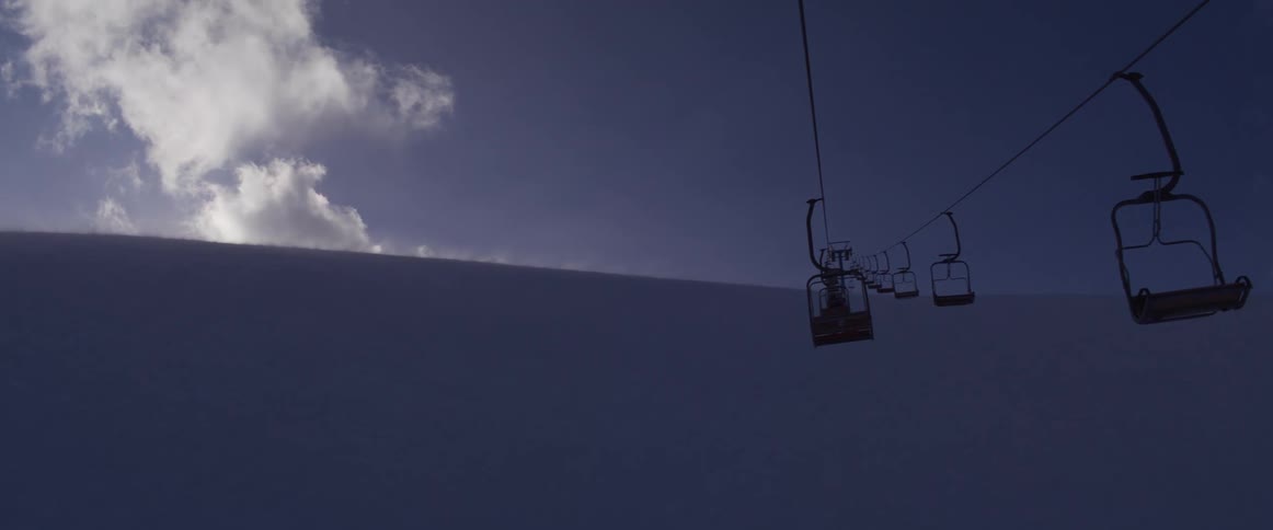 A stationary ski lift hangs over a vast, snow-covered mountain under a moody blue sky with passing clouds