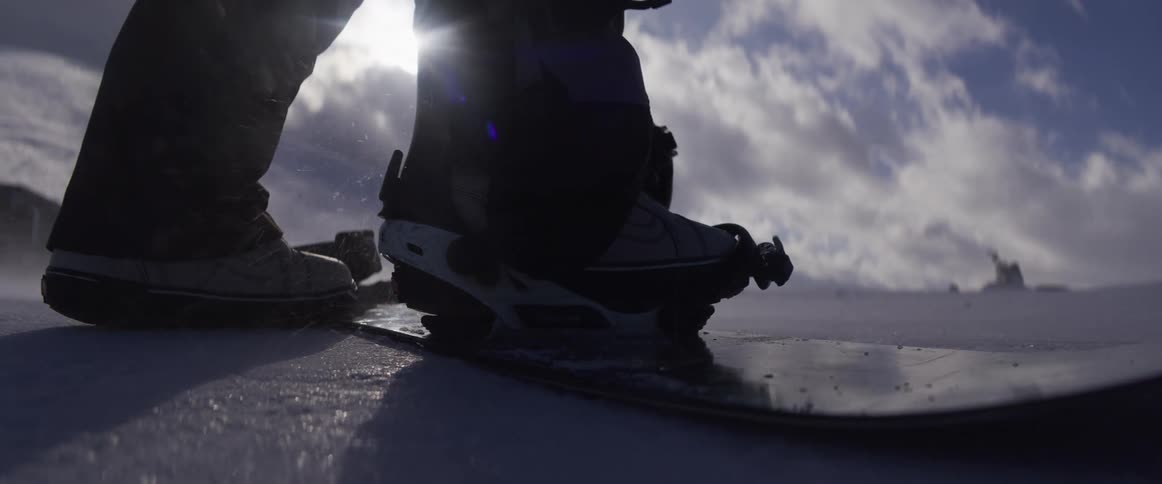 A snowboarder straps into their bindings in slow motion, backlit by the bright winter sun on a snowy mountain
