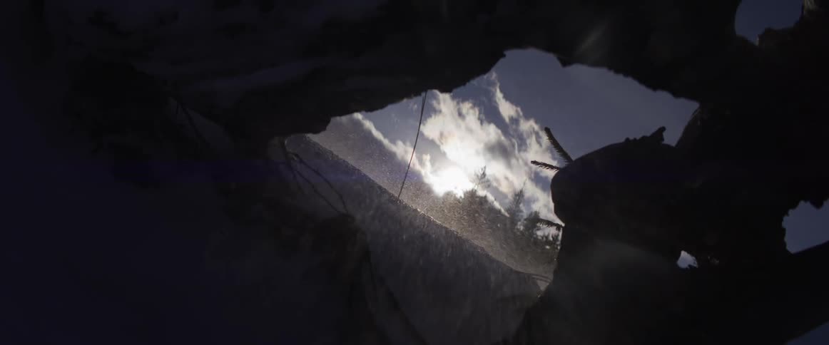 A windy, snowy mountain peak is seen through an opening in an ice cave as the sun shines brightly in the sky