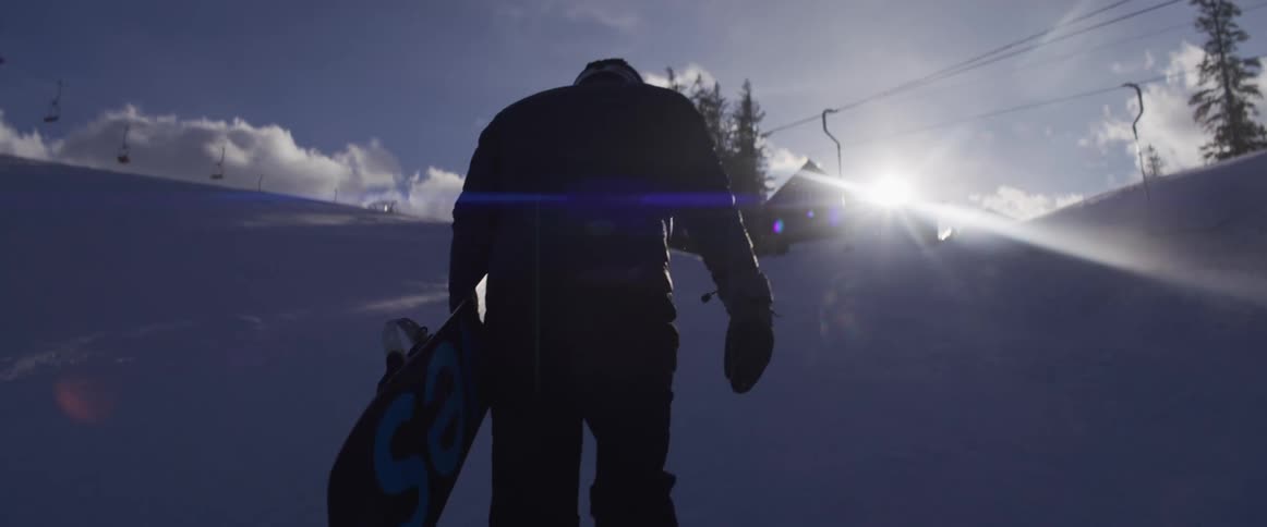A silhouetted snowboarder in a helmet walks up a snowy mountain slope carrying their board towards the bright sun in slow motion