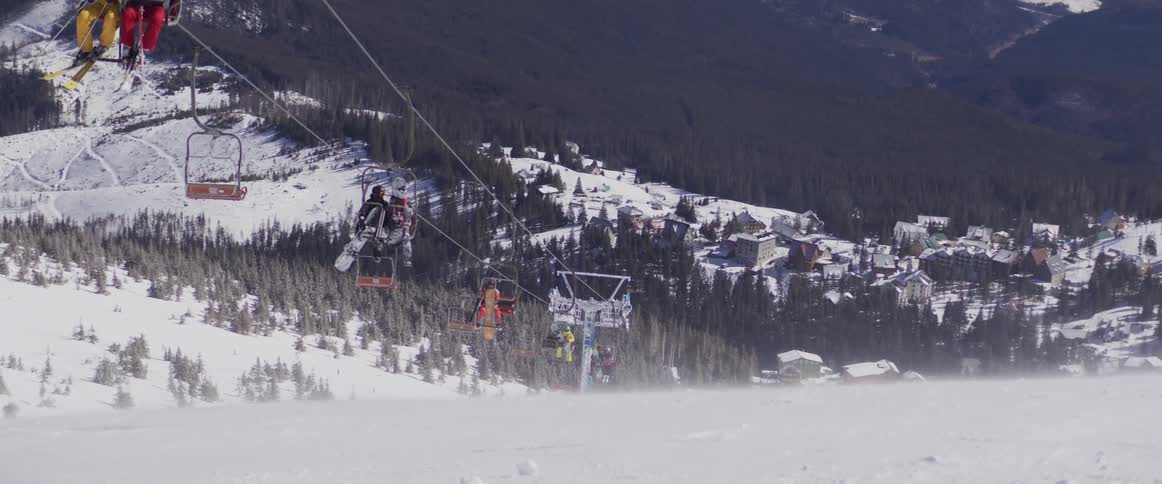 Skiers and snowboarders ride a chairlift over a windy, snow-covered slope at a mountain ski resort
