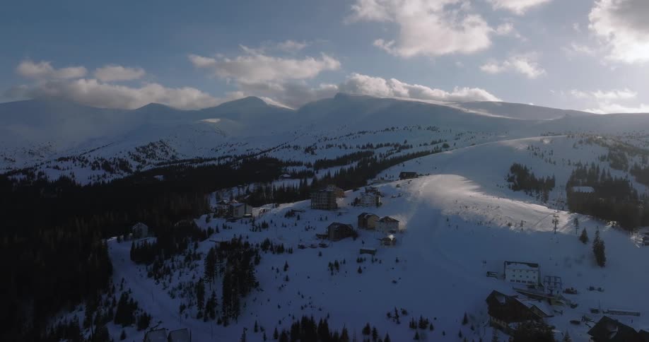 An aerial view of a remote ski resort village on a snow-covered mountain with sunlight casting shadows across the slopes
