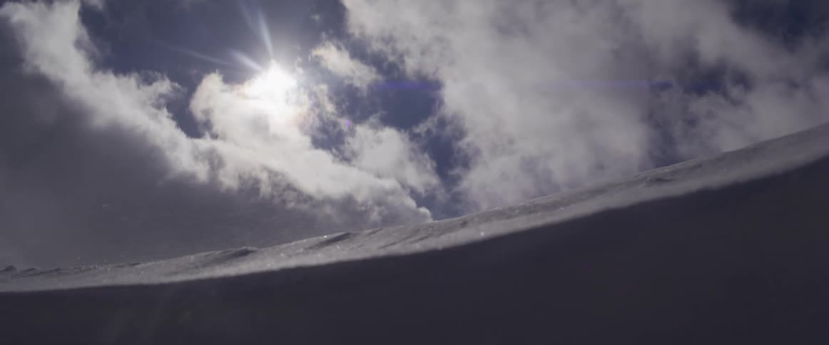 Slow motion of a powerful blizzard blowing snow across a mountain peak as the sun shines brightly through the clouds
