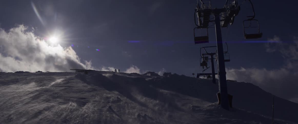 A ski lift stands silhouetted against the bright sun as fierce winds whip snow across a mountain peak
