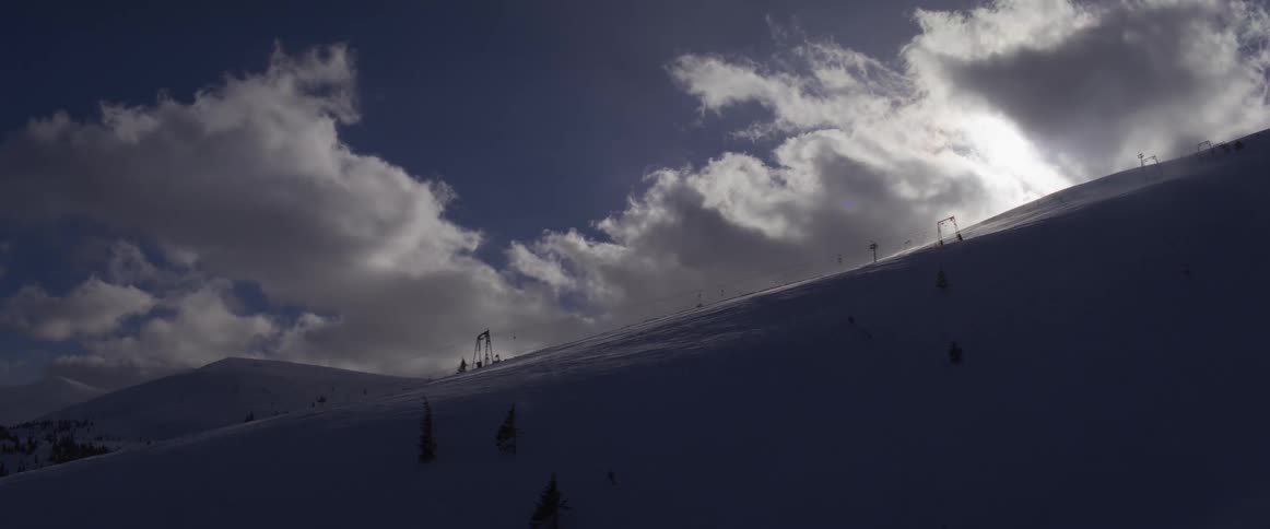 A silhouetted ski lift on a snowy mountain is back-lit by the sun breaking through the clouds