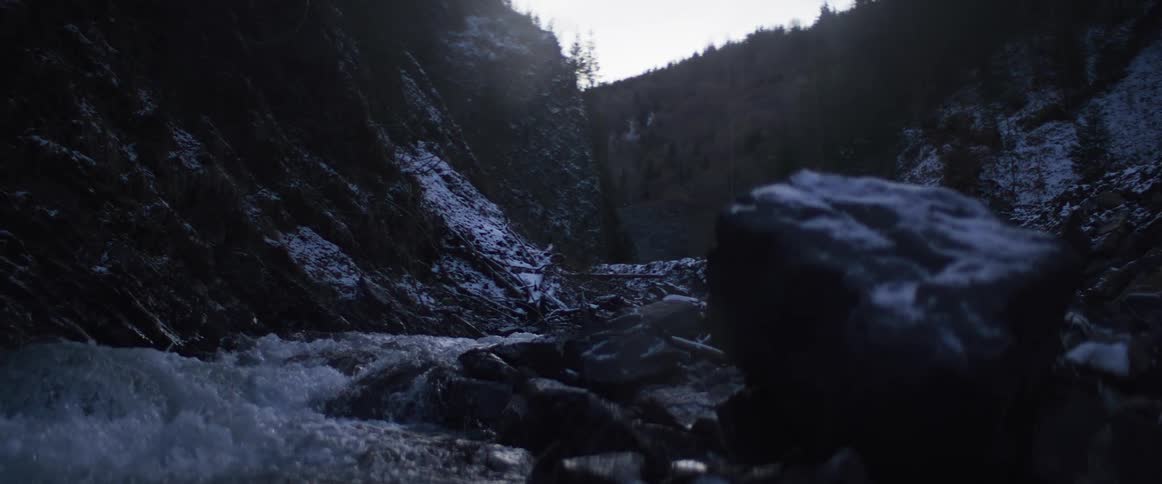 A cold winter river rushes through a snowy, rocky gorge at twilight, with a rack focus from a foreground rock to the background