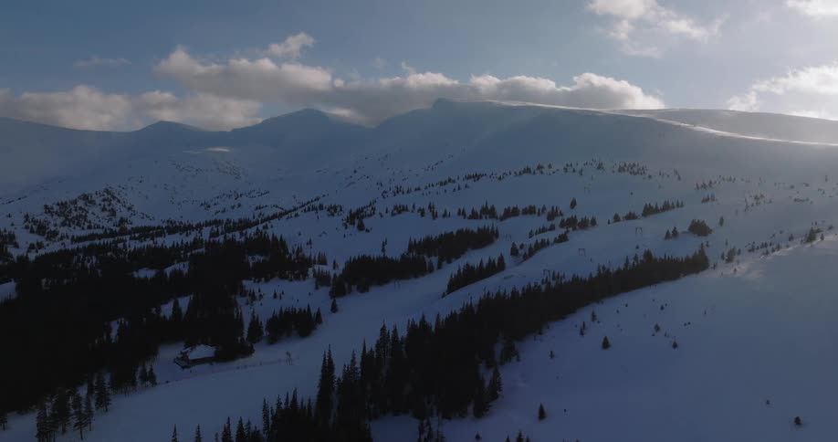 A majestic aerial pull-back shot reveals a sprawling snow-covered mountain range and ski resort bathed in the golden light of a winter day