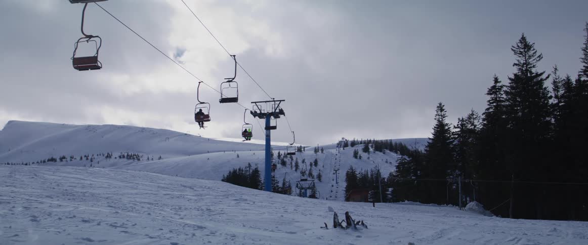 Skiers ride a chairlift up a snowy mountain at a ski resort on an overcast winter day