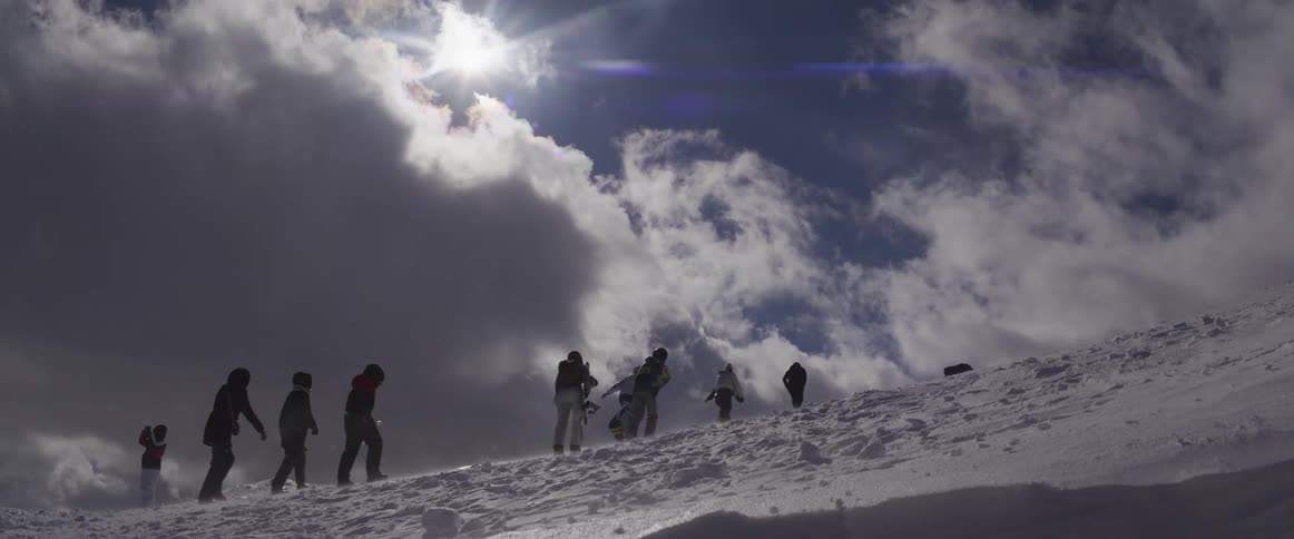 Silhouetted group of snowboarders and hikers ascend a windy, snow-covered mountain peak against a dramatic, sunlit sky in slow motion