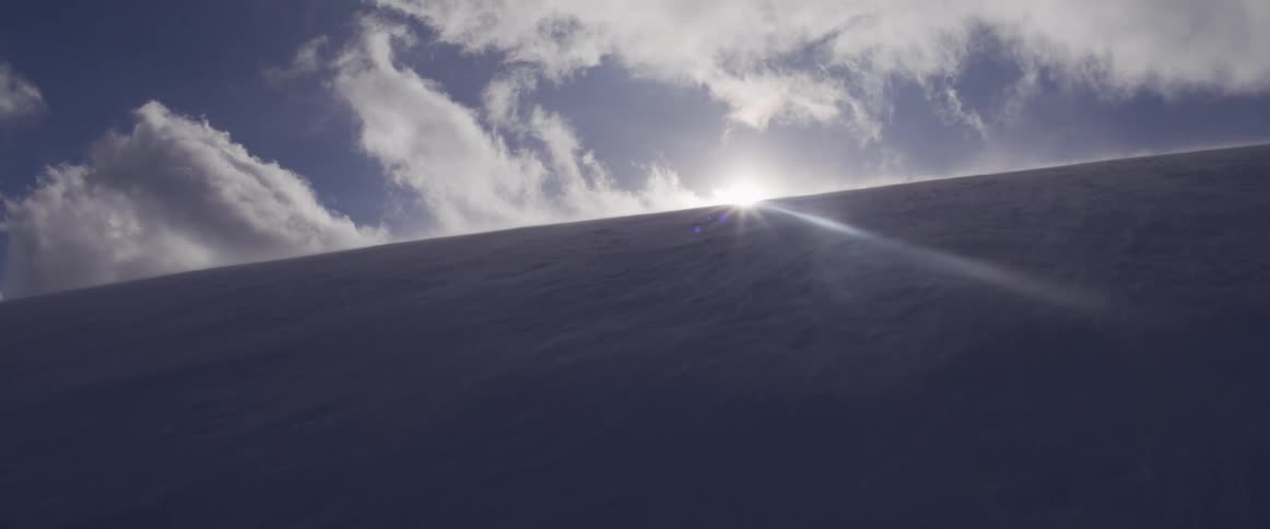 Sunlight slowly emerges over a snowy, wind-blasted mountain ridge, creating a dramatic lens flare