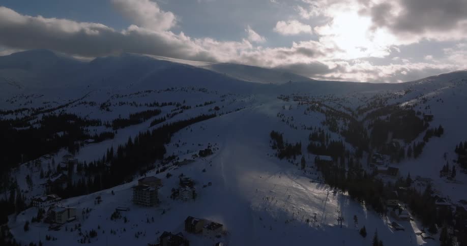 Aerial view slowly pulls back over a snowy mountain ski resort under a bright, cloudy sky