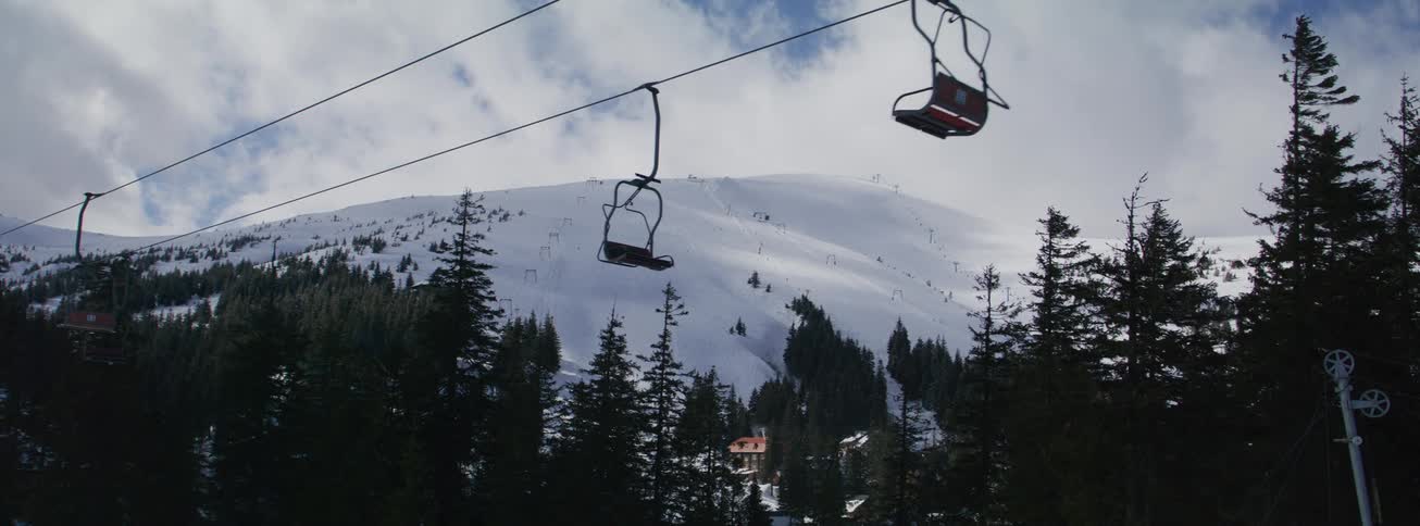 Empty chairlifts glide slowly over pine trees towards a vast, snowy ski mountain on a cloudy day