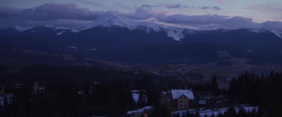 A snow-capped mountain peak looms over a quiet, snowy village in the valley during a cold winter twilight