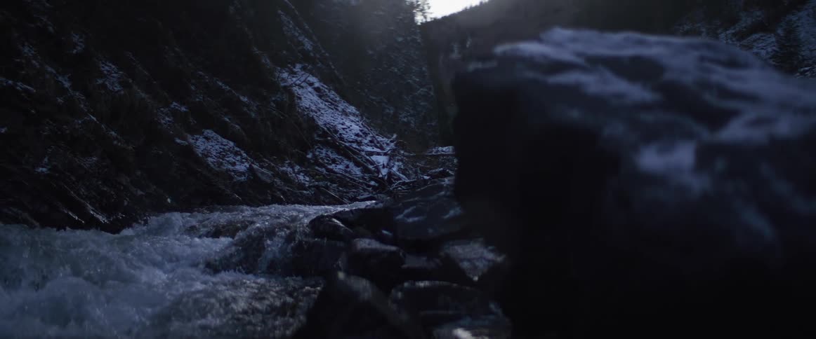 Focus pulls from a dark rock to reveal a powerful river flowing through a remote, snowy canyon