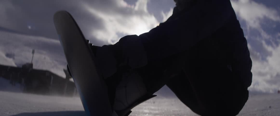 Slow motion close up of a snowboarder strapping into their board on a windy mountain top, backlit by the bright sun before riding away
