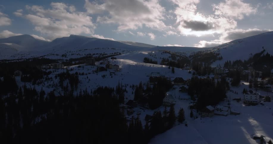 Slow aerial ascent reveals a tranquil, snow-covered mountain ski resort village under a cloudy, sunlit sky