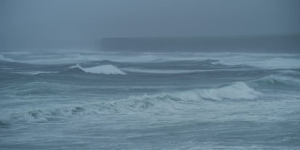 A dramatic slow-motion shot captures the raw power of the ocean as large waves churn during a heavy storm with dense fog