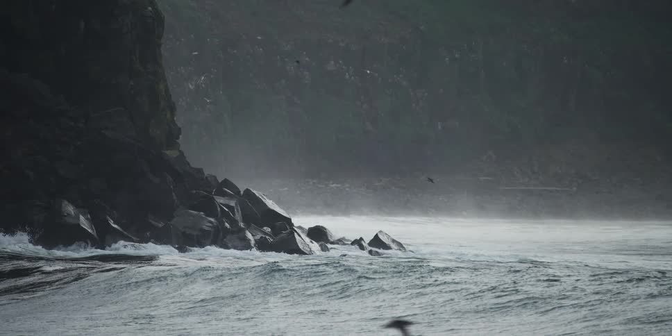Dramatic slow motion of powerful ocean waves crashing against a rocky coastline as seabirds fly through the mist