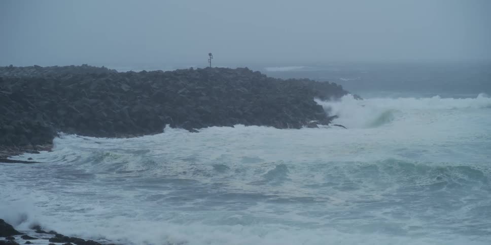 Slow motion footage shows powerful storm waves crashing against a rocky breakwater on a grey, foggy day