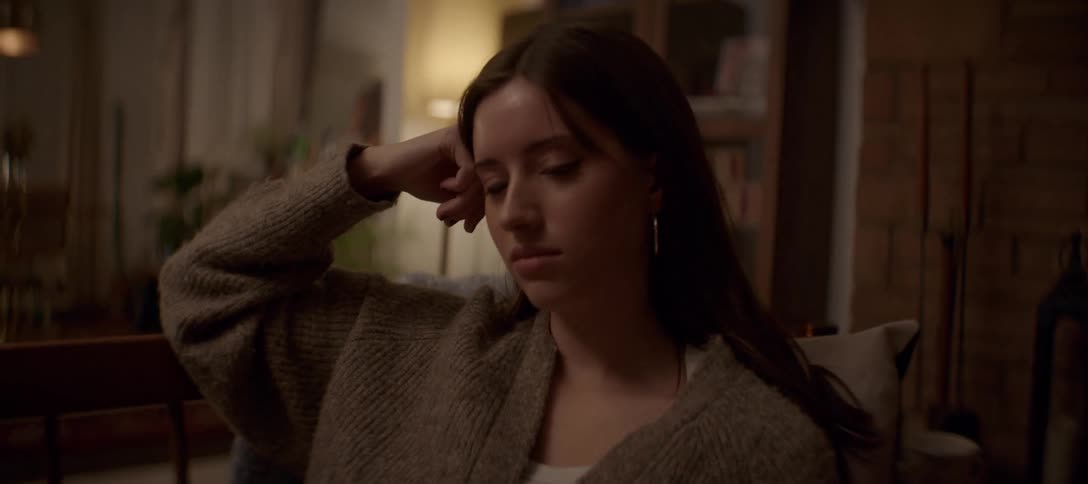 A thoughtful young woman in a cozy sweater rests her head in her hand while reading a book in a warm, dimly lit room