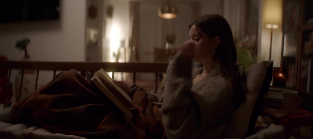 A relaxed woman enjoys a cozy night in, reading a book while curled up on the sofa with a warm blanket in her living room