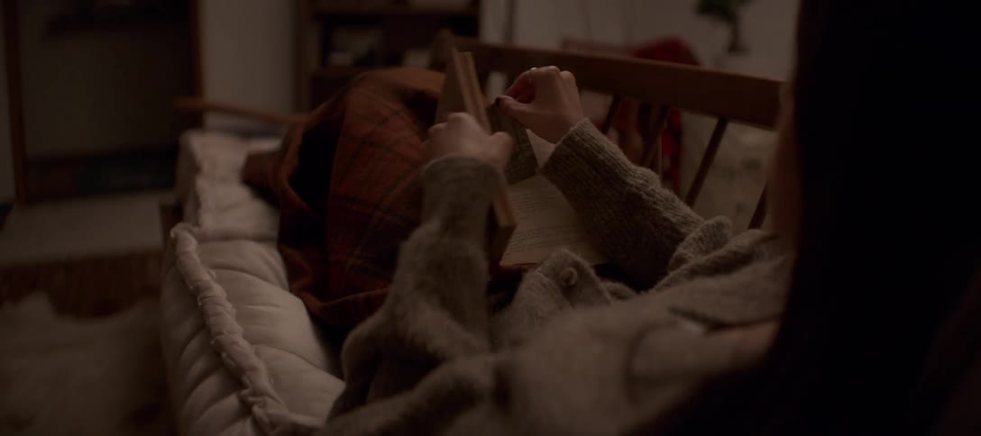 A woman in a cozy sweater relaxes on a chaise lounge, reading a book in a dimly lit room before closing it