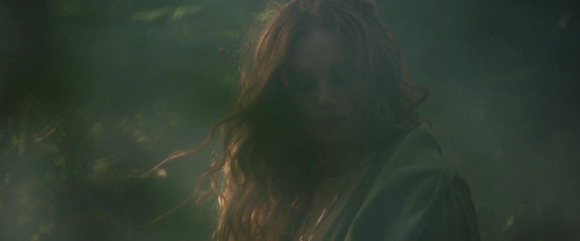 A young woman with long curly hair and a gray cloak sits in the fog
