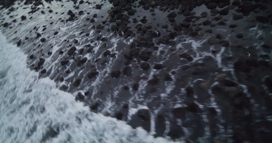 A dynamic aerial view flies over a dark, rocky shoreline as foamy waves crash, eventually pulling back to reveal the vast, churning ocean