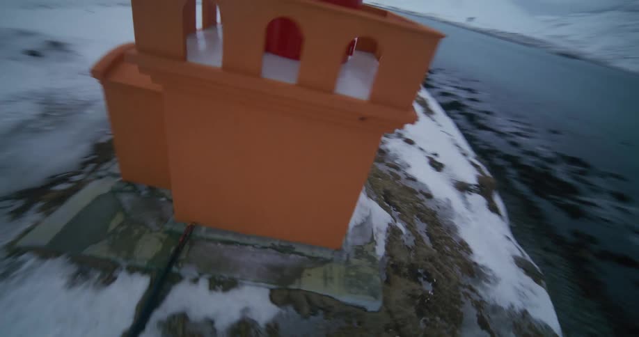 An FPV drone speeds across a snowy Icelandic landscape, flying towards and circling a vibrant orange lighthouse on the coast