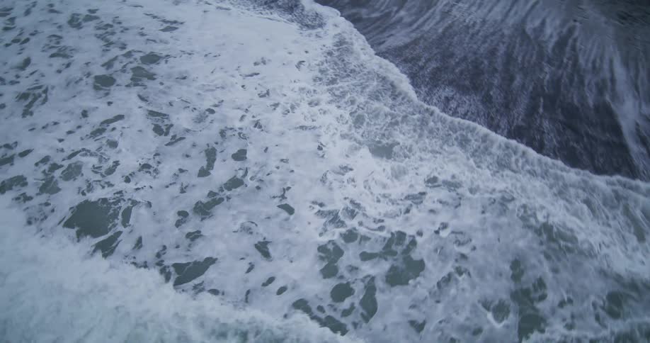 A mesmerizing slow-motion aerial view of foamy ocean waves rolling onto a dark sand beach
