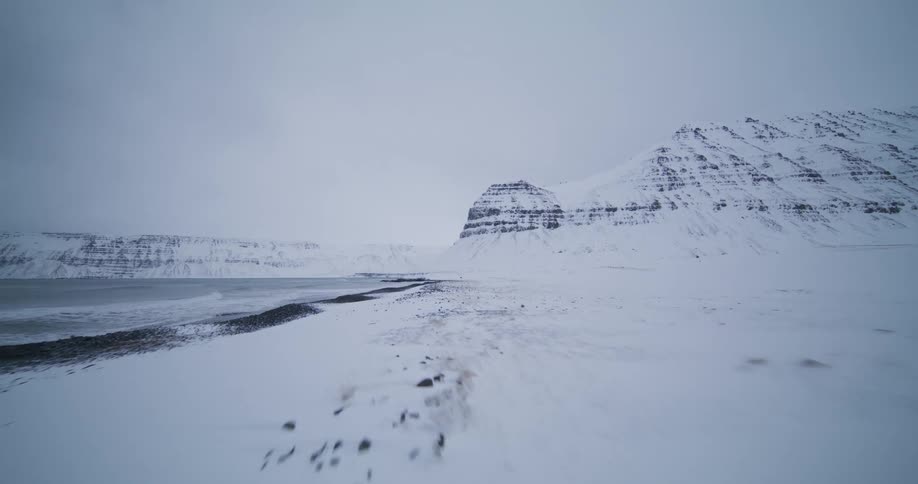 A breathtaking low aerial shot glides over a snow-covered beach along a majestic, mountainous coastline in winter