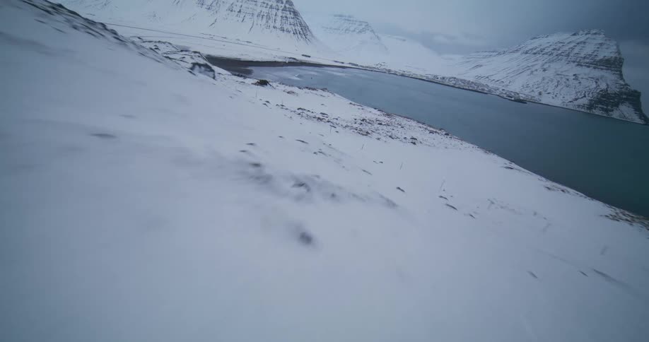 A dynamic FPV drone shot races down a vast, snow-covered hillside, revealing a frigid Icelandic fjord surrounded by mountains