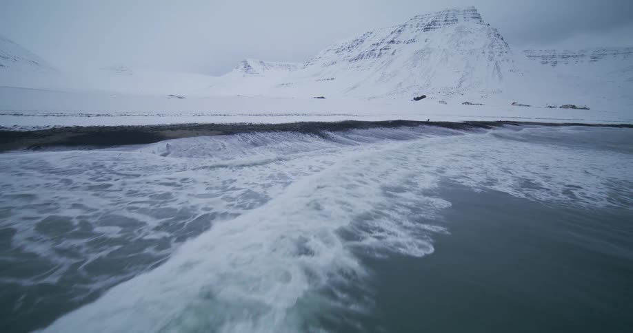 A powerful ocean wave breaks in slow motion onto a snowy beach with majestic snow-covered mountains in the background