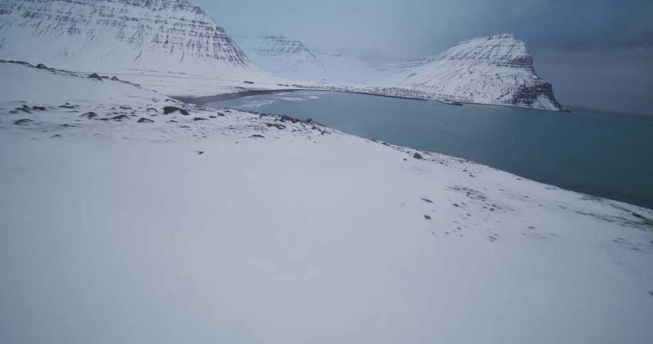 A fast-paced aerial shot flies over a snowy Icelandic mountain to reveal a majestic fjord and a remote coastal village