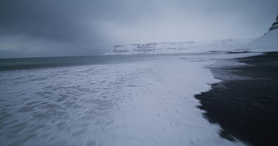 A fast-moving aerial shot flies low over a snow-covered beach and into the icy waves of a cold ocean