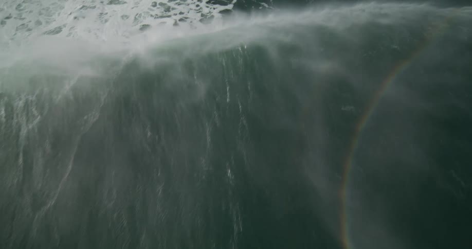 A mesmerizing high-angle slow-motion shot captures the raw power of a giant ocean wave with a rainbow forming in its spray