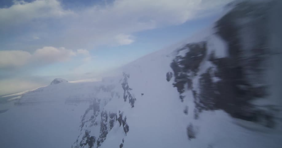 A fast-paced first-person aerial view flying along the precipice of a vast, snow-covered mountain