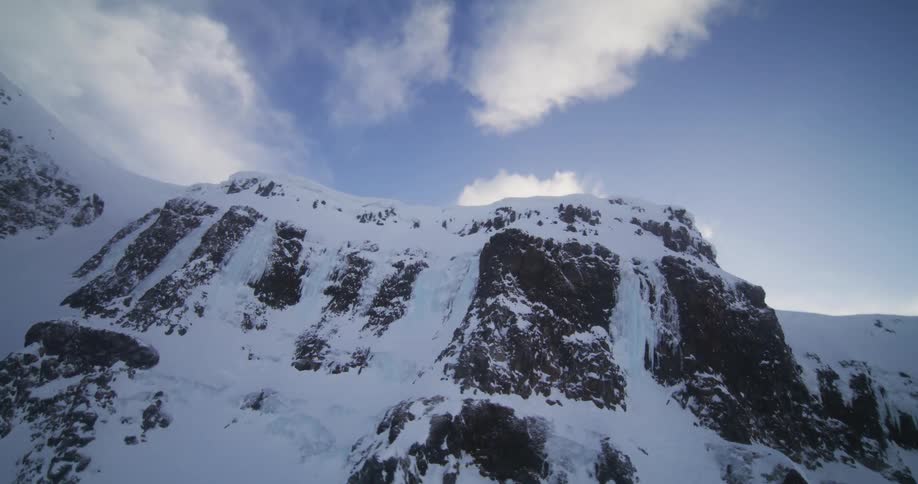 An aerial view pulls back from a steep, icy mountain face to reveal a vast, snow-covered landscape under a partly cloudy sky