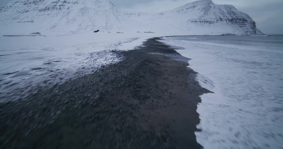A dramatic aerial shot flies along a desolate, snow-covered coastline, revealing towering mountains and dark waves crashing onto a black sand beach