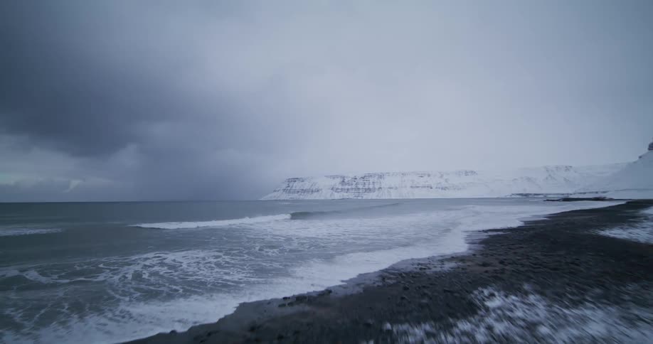 A fast-paced aerial shot glides over a snow-covered beach towards the ocean on a cold, overcast day