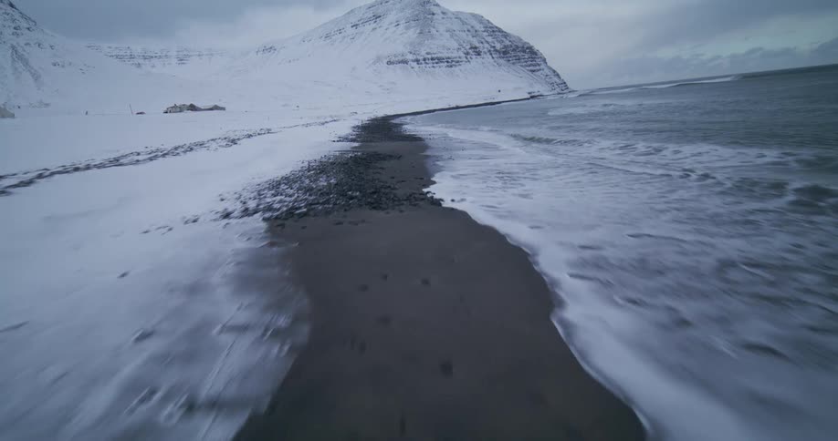 A thrilling low-altitude aerial shot speeds along a desolate, snow-covered coastline where dark sand meets the cold ocean waves