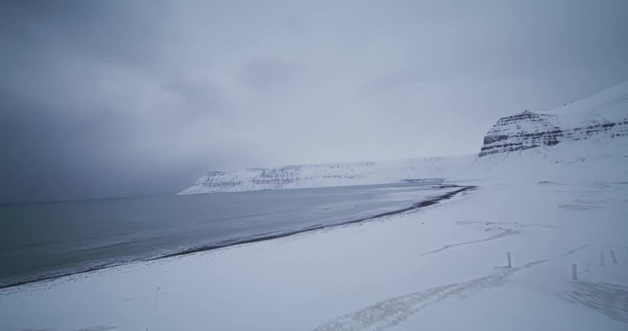 An aerial view flies over a solitary white church with a red roof on a snow-covered beach next to the ocean on a stormy winter day
