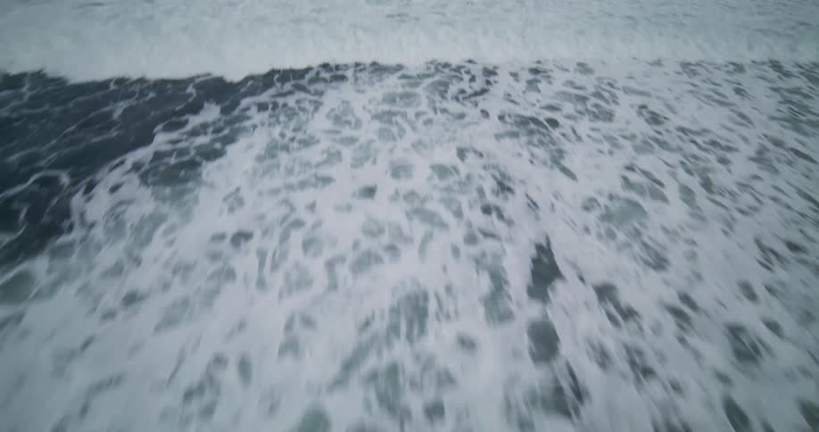 A fast-paced aerial flyover from a dark rocky shore into the churning white foam of the ocean waves