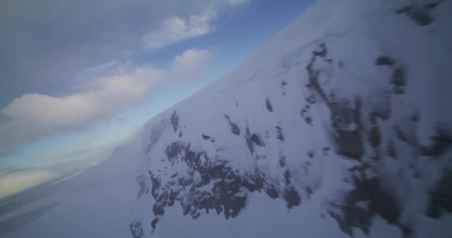 A thrilling first-person aerial view flying alongside the edge of a vast, snow-covered mountain cliff under a cloudy blue sky